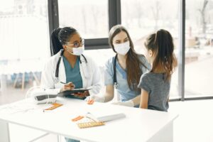 Two female doctors in masks assist a child in a bright medical room.