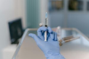 Close-up of a blue gloved hand holding a medical syringe in a clinical setting.